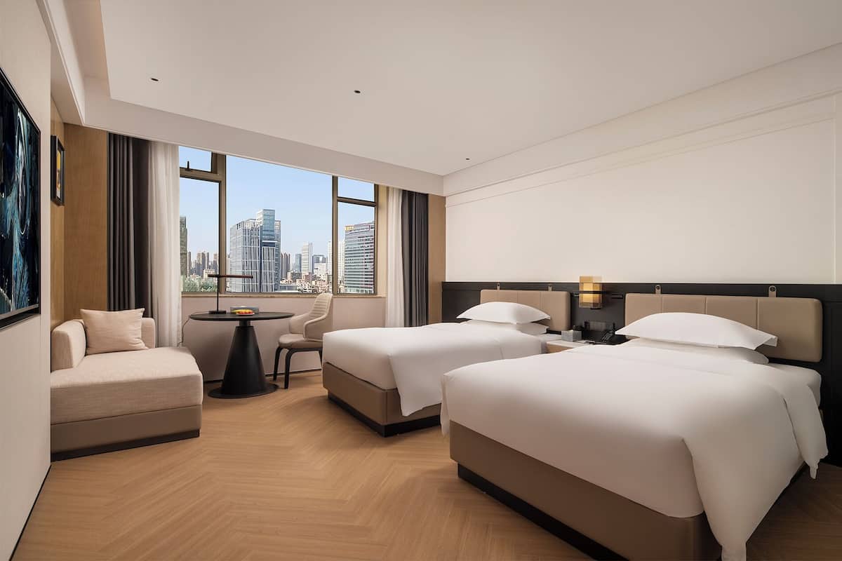 Habitación de hotel con dos camas, un sofá, una mesita y grandes ventanales con vistas a la ciudad.