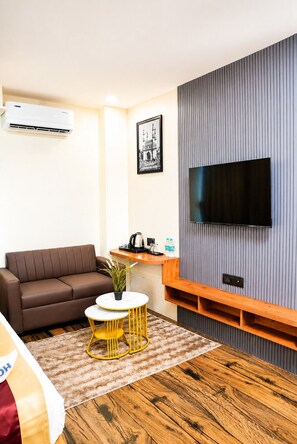 Una sala moderna con un sofá marrón, una mesita redonda pequeña, un televisor montado en una pared gris y un estante de madera debajo.