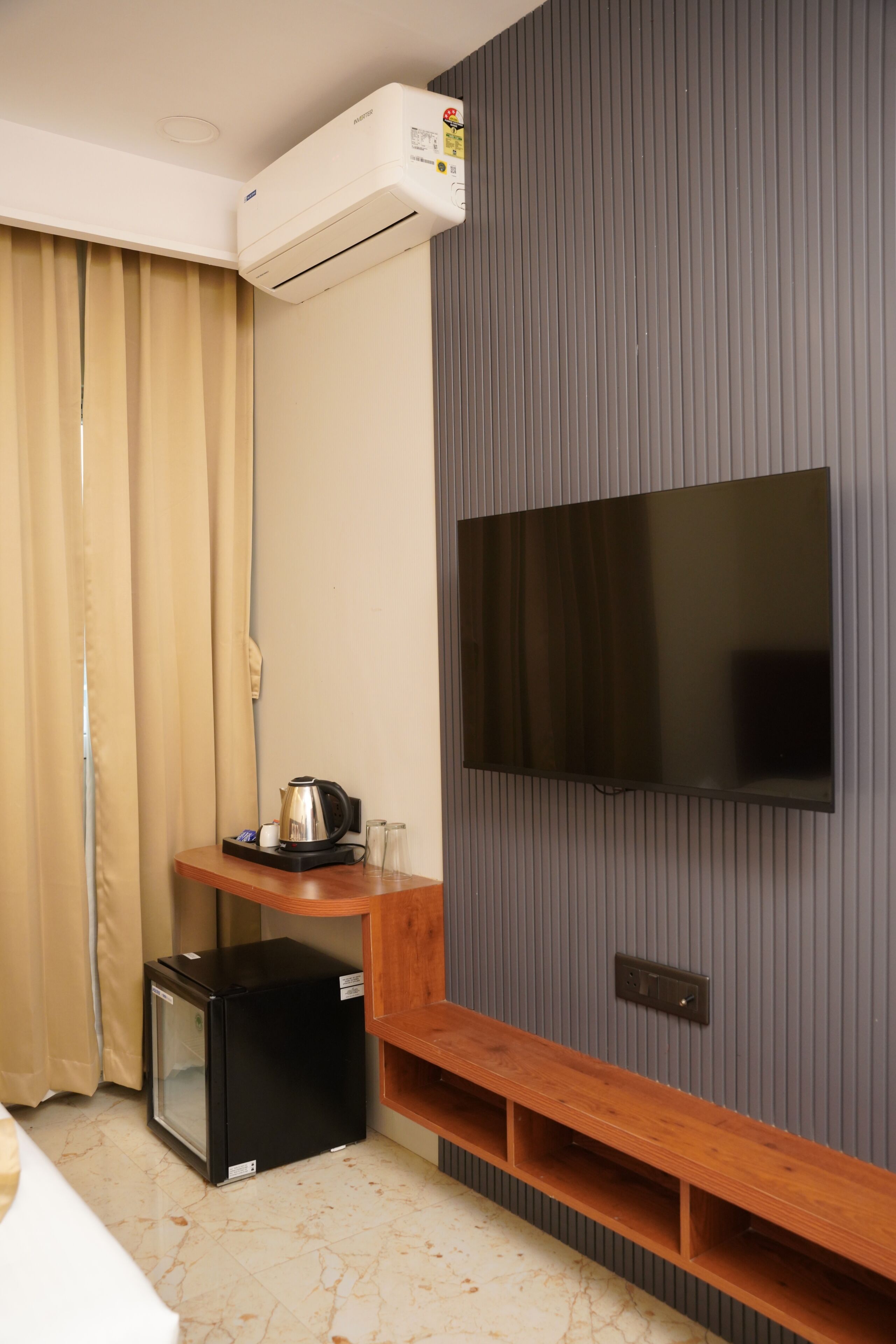 Habitación de hotel con televisor de pantalla plana montado en una pared gris, una mesita de madera con una tetera, un refrigerador pequeño y una ventana con cortinas.
