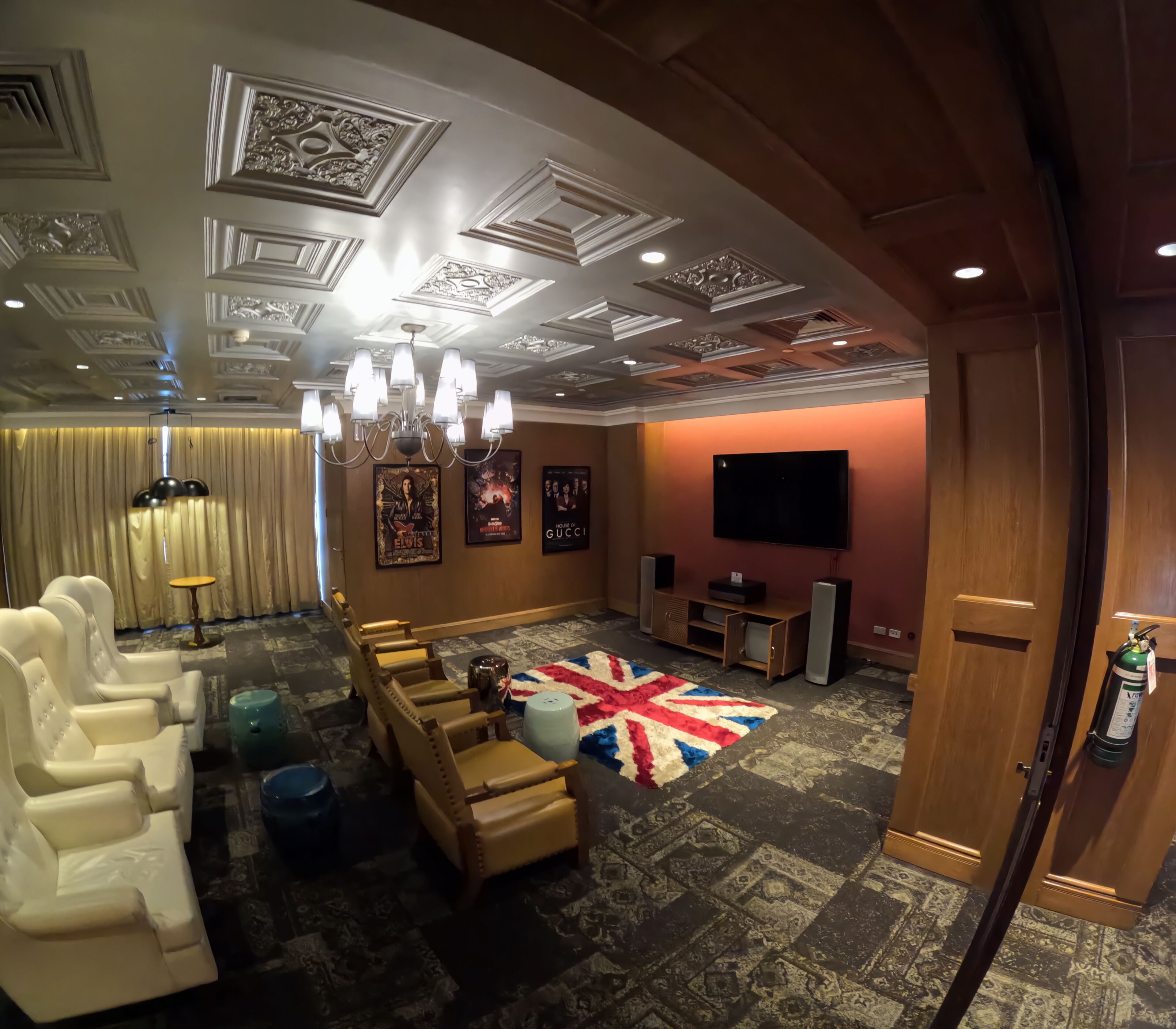 A room with a ceiling with intricate designs, a chandelier, a flat-screen TV, a Union Jack rug, and a row of sofas.