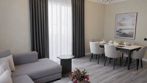 Un salon moderne avec un canapé gris, une table basse ronde ornée d’un vase de fleurs, un coin repas avec une table dressée pour quatre personnes, et une grande fenêtre avec des rideaux légers.