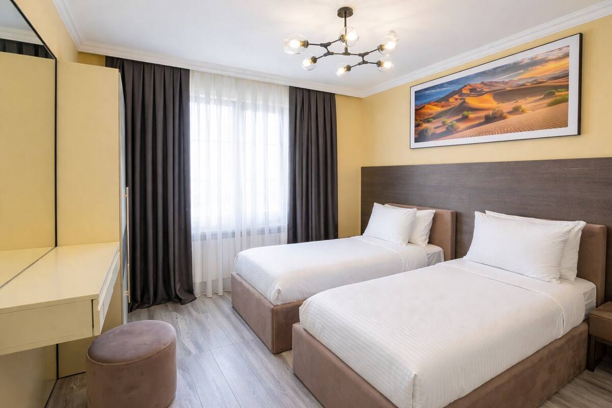 A hotel room with two beds, a desk, a chair, a large window with curtains, and a painting of a desert landscape.