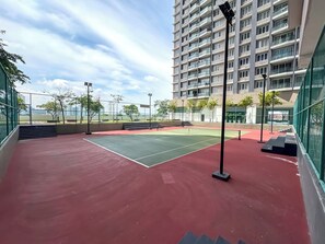 An outdoor tennis court with a red surface, surrounded by a fence, and a high-rise building in the background. - Aurora Residence by Manhattan Group (Puchong)