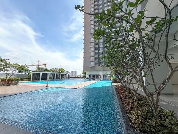 A swimming pool with tiled flooring, surrounded by greenery and a tall building in the background. - Aurora Residence by Manhattan Group (Puchong)