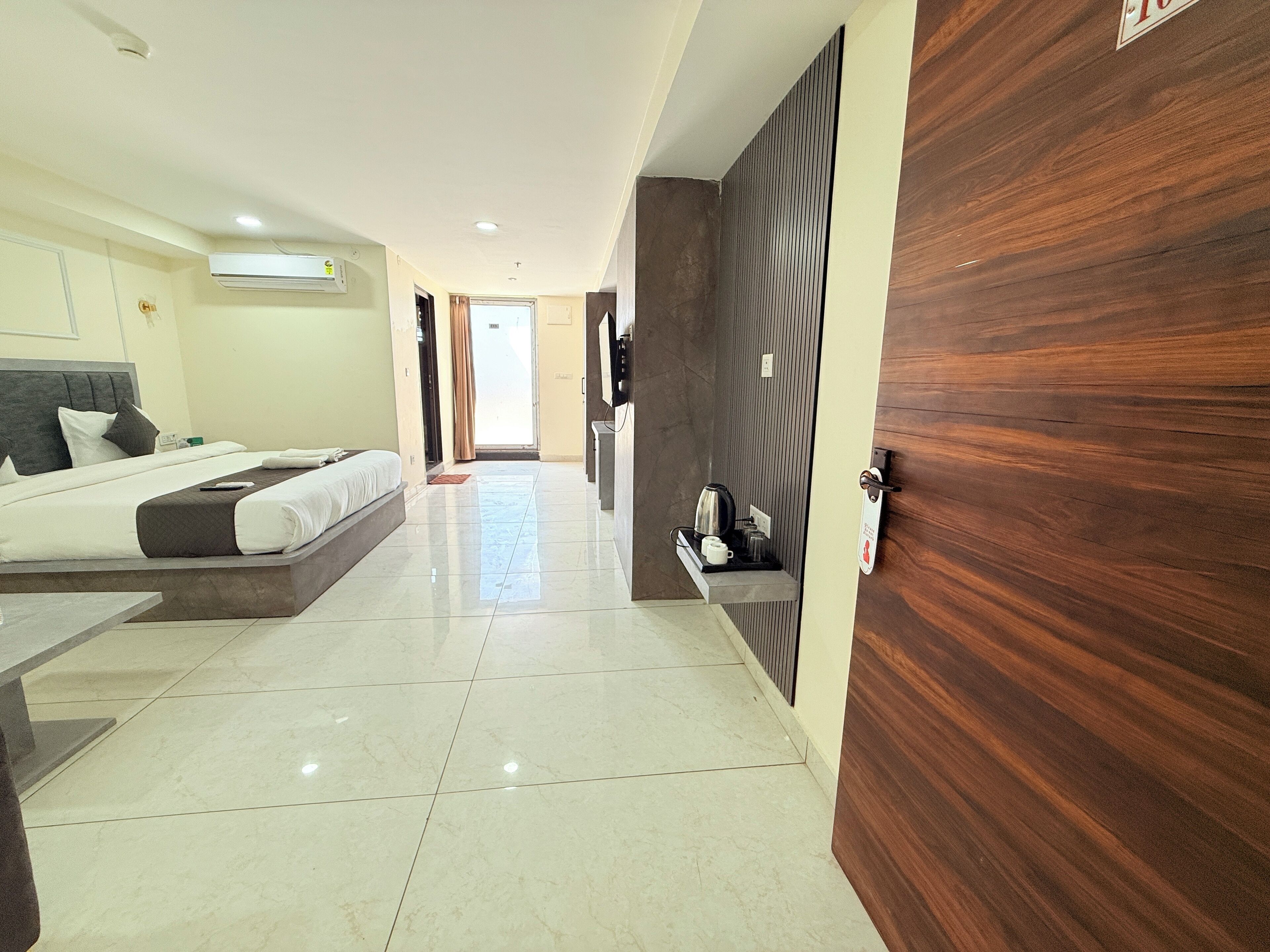 A modern hotel room with a bed, a small desk, a wooden panel wall, and a view of the hallway.