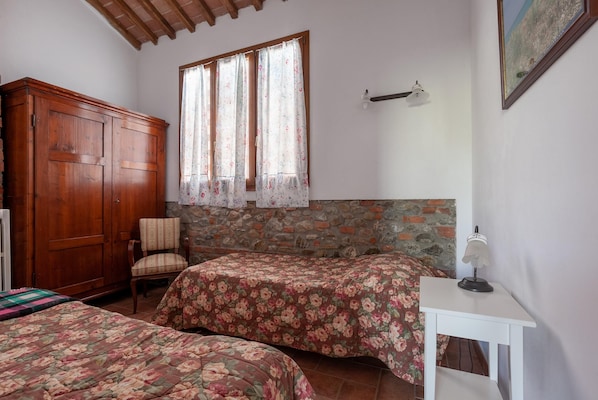 Ein Schlafzimmer mit zwei Betten, einem Holzschrank, einem Stuhl, einem kleinen Tisch mit Lampe und einem Fenster mit Vorhängen.