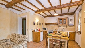 A cozy kitchen with wooden beams, a dining table with chairs, a floral sofa, and a small refrigerator. - A Cottage Wrapped in Cypress Dreams (Lucignano)