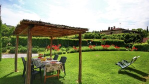 A wooden pergola with a table set for dining, surrounded by lush greenery and red flowers. - A Cottage Wrapped in Cypress Dreams (Lucignano)