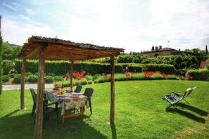 Una pergola in legno con un tavolo apparecchiato per mangiare, circondata da vegetazione lussureggiante e fiori rossi.