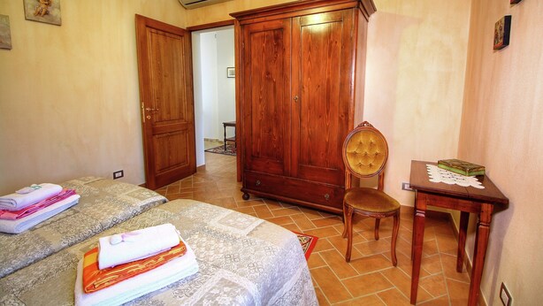 A bedroom with two beds, a wooden wardrobe, a chair, and a small table with a book. - A Cottage Wrapped in Cypress Dreams (Lucignano)
