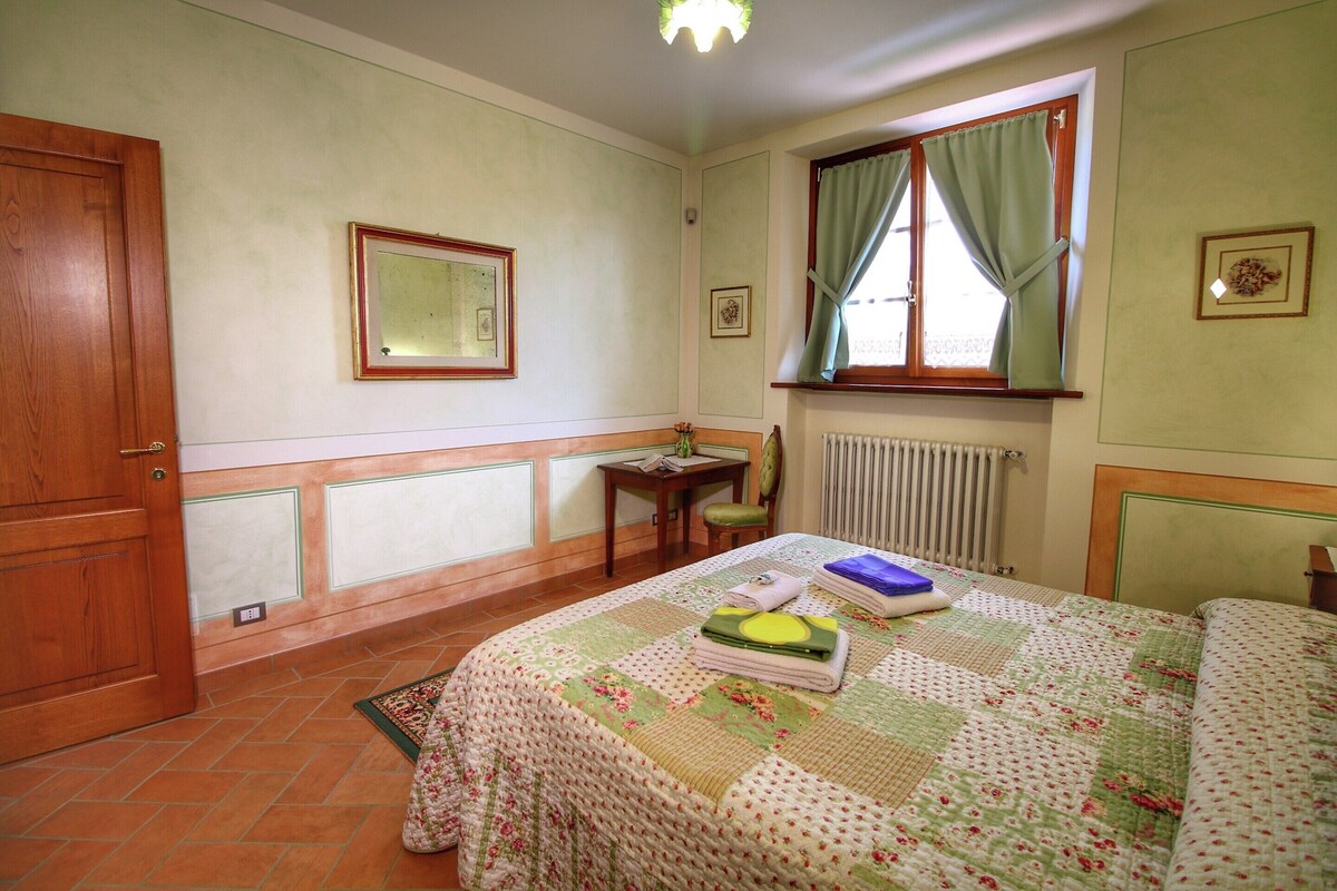 Ein Schlafzimmer mit Bett, Holztür, kleinem Tisch mit Stuhl, Fenster mit Vorhängen und Heizkörper.