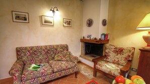 A living room with a floral sofa, a wooden armchair, a fireplace, and a fruit bowl on a table. - A Cottage Wrapped in Cypress Dreams (Lucignano)