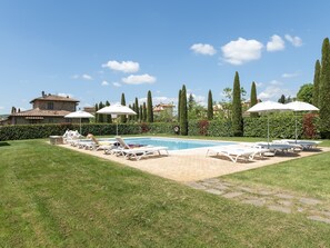 Area piscina con lettini e ombrelloni, circondata da siepi e alberi alti.