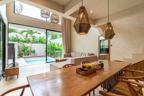 A modern living room with a pool, a dining area with a wooden table and chairs, and a large window with a view of greenery. - Serene Villas Bingin, Uluwatu by Blacksand (Pecatu)