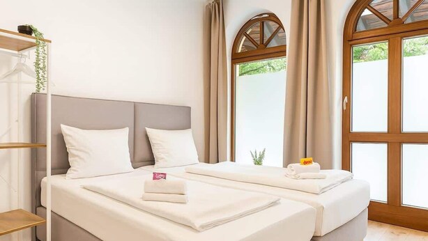 Una cama doble con sábanas y almohadas blancas, una mesita de noche con una toalla y una planta pequeña, y una ventana con cortinas.