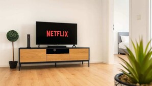 Una sala moderna con un televisor de pantalla plana mostrando el logotipo de Netflix, un mueble de madera, una pequeña planta en maceta y una vista parcial de un dormitorio al fondo.