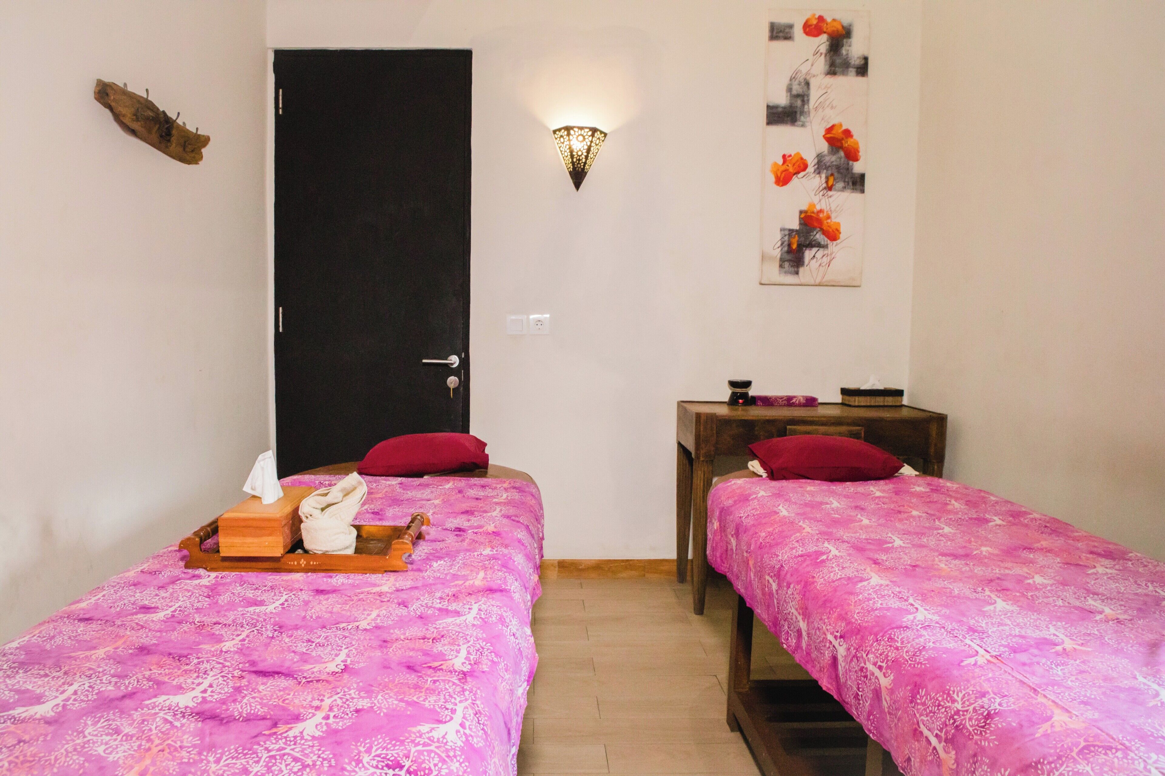 A room with two beds, each with a pink patterned bedspread, a wooden nightstand with a lamp, and a wall-mounted artwork.