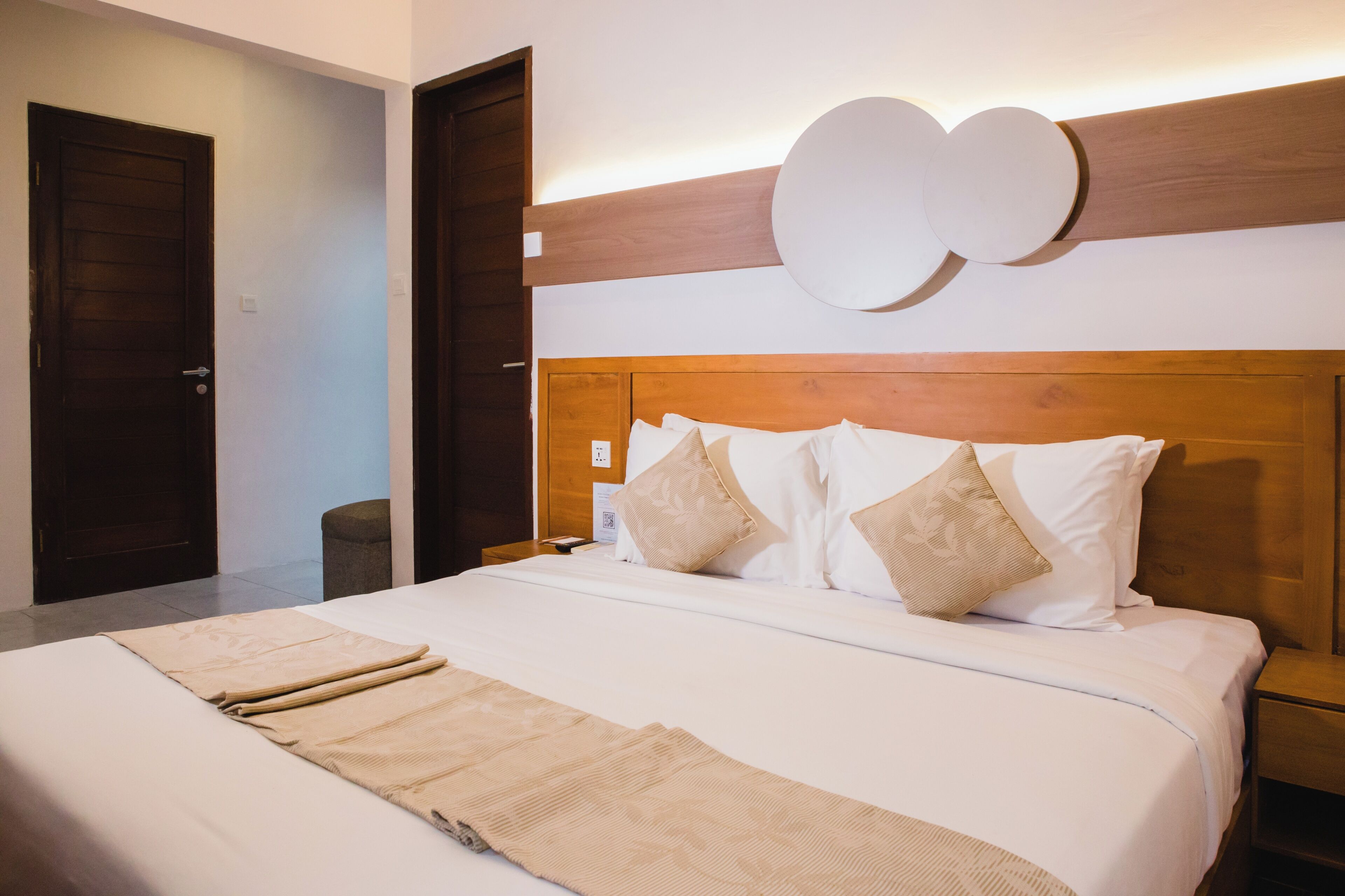 A neatly made bed with white linens and beige throw pillows, a wooden headboard, and a wall-mounted circular light fixture.