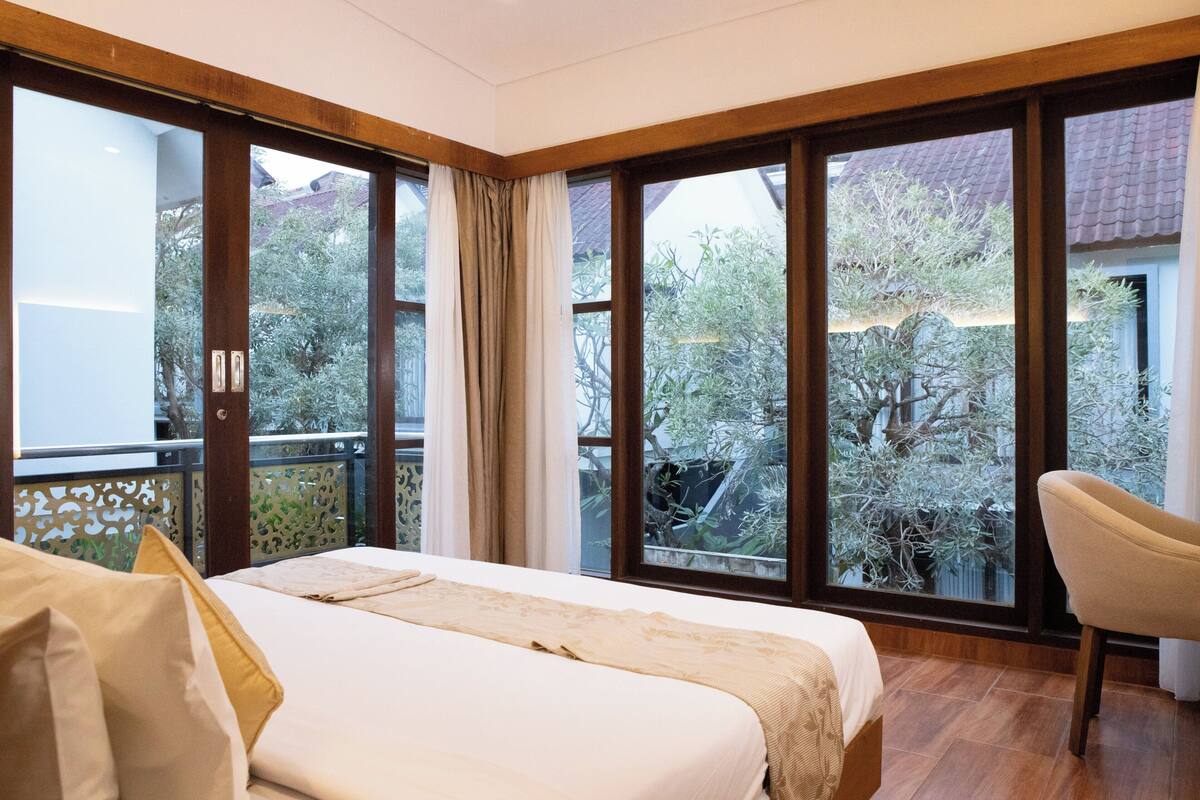 Une chambre d’hôtel avec un grand lit, une chaise et un balcon donnant sur des arbres et des immeubles.