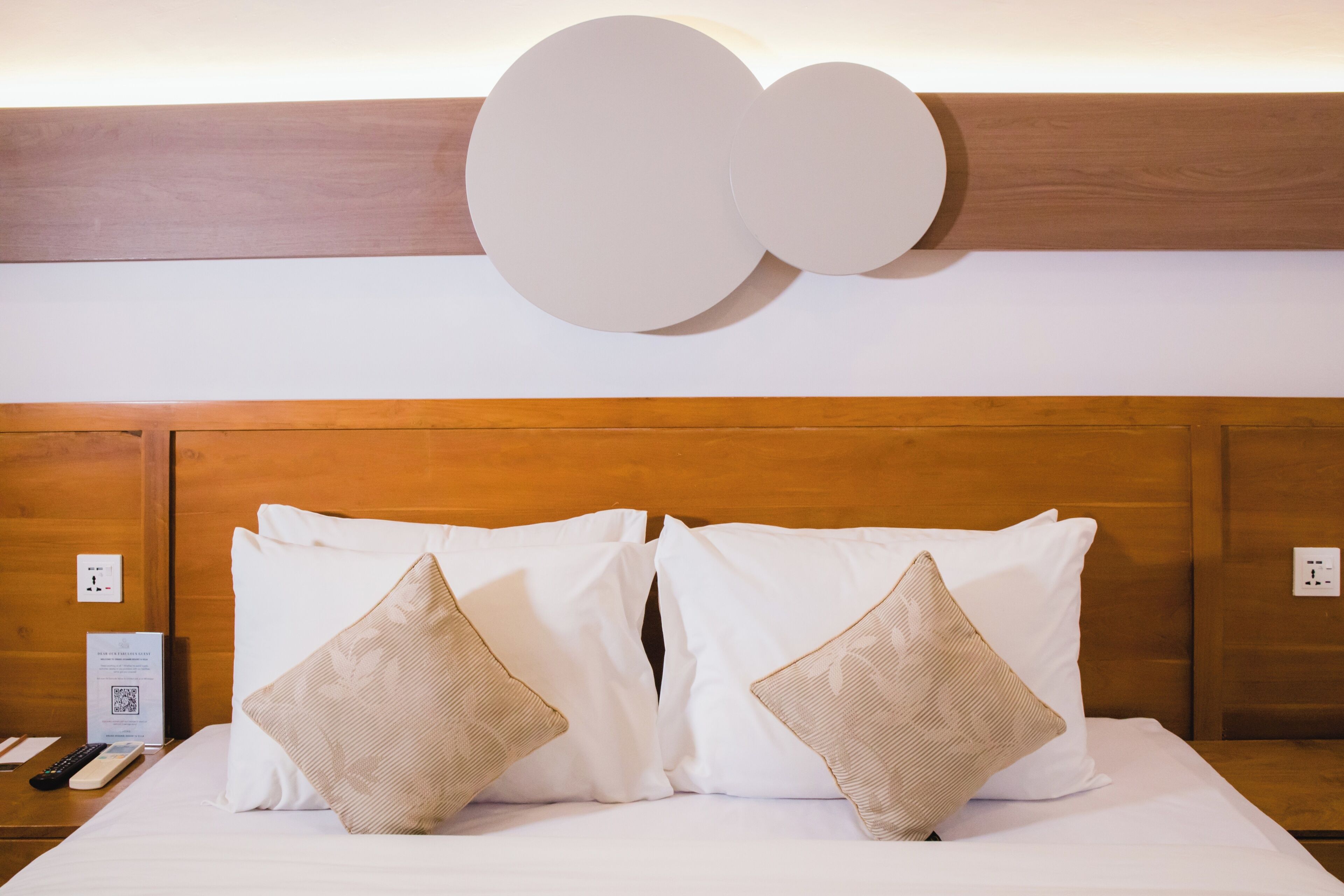 Une chambre d’hôtel avec une tête de lit en bois, deux oreillers blancs et un luminaire fixé au mur.