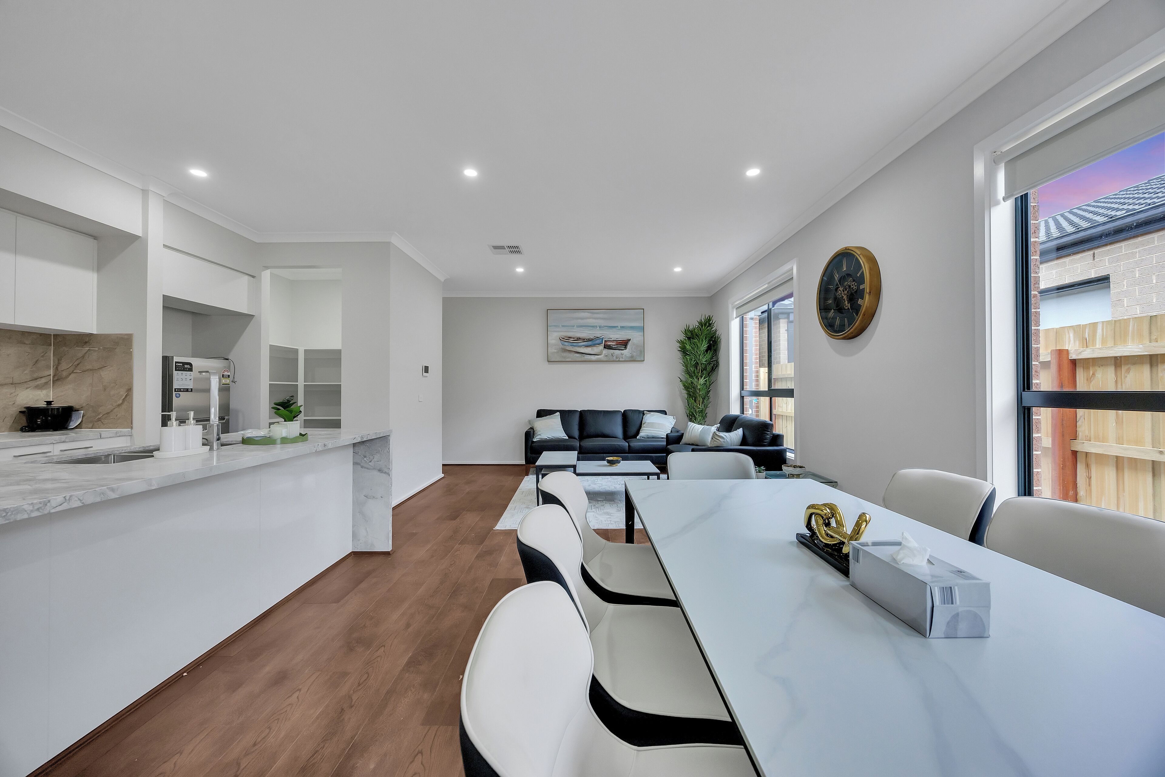 A modern kitchen with a marble countertop, a built-in oven, and a sink. There is a dining area with a white table and chairs, a plant, and a painting on the wall.