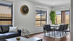En moderne stue med sofa, sofabord, spisebord med stole, et stort ur og udsigt til baghaven gennem store vinduer.