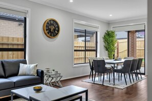 Una sala de estar moderna con sofá, mesa de centro, mesa de comedor con sillas, un reloj grande y vistas del jardín trasero a través de amplias ventanas.