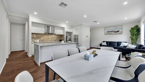 Et moderne køkken med hvide skabe og en marmor bordplade, et spiseområde med et hvidt bord og stole, og en opholdsstue med en sort sofa og et maleri af både.