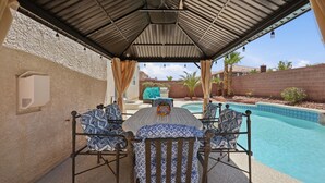 A covered outdoor seating area with a pool, featuring a table and chairs with patterned cushions, a pergola overhead, and a view of the pool and surrounding landscape. - Vegas Grand Villa - 5BR, Pool, Putting Green (Las Vegas)