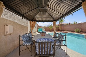 Ein überdachter Außenbereich mit Pool, ausgestattet mit einem Tisch und Stühlen mit gemusterten Kissen, einer Pergola und Blick auf den Pool und die umliegende Landschaft.