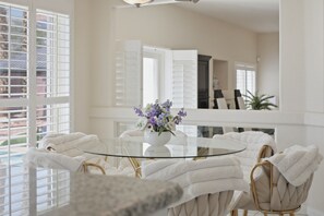 A room with a round glass table surrounded by white upholstered chairs, a white vase with purple flowers, and a view of the outside through large windows with white shutters. - Vegas Grand Villa - 5BR, Pool, Putting Green (Las Vegas)