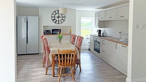 A modern kitchen with white cabinets, a wooden dining table with plaid chairs, a clock on the wall, and a view of the outdoors. - 3 bedroom accommodation in Brough (Brough)