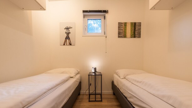 A small, well-lit room with two single beds, a small table with a lamp, a window, and a wall-mounted picture. - Drenthe Wellness Lodge for 4 (Ruinen)