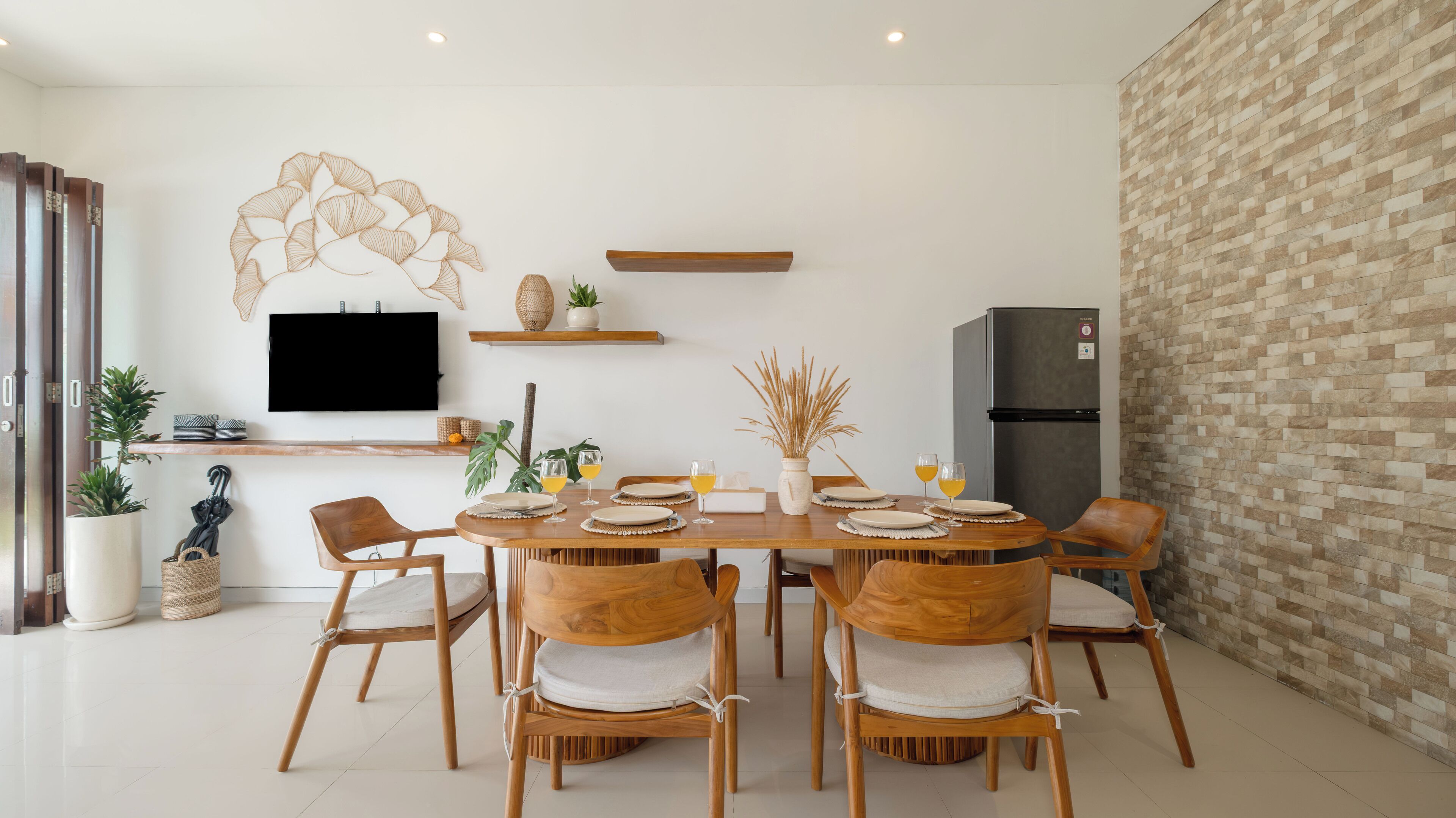 Ein moderner Essbereich mit einem Holztisch, gedeckt für sechs Personen, umgeben von Korbstühlen mit weißen Kissen. Ein Flachbildfernseher ist an der Wand über dem Tisch angebracht, und es gibt Regale mit Dekoration und Pflanzen.