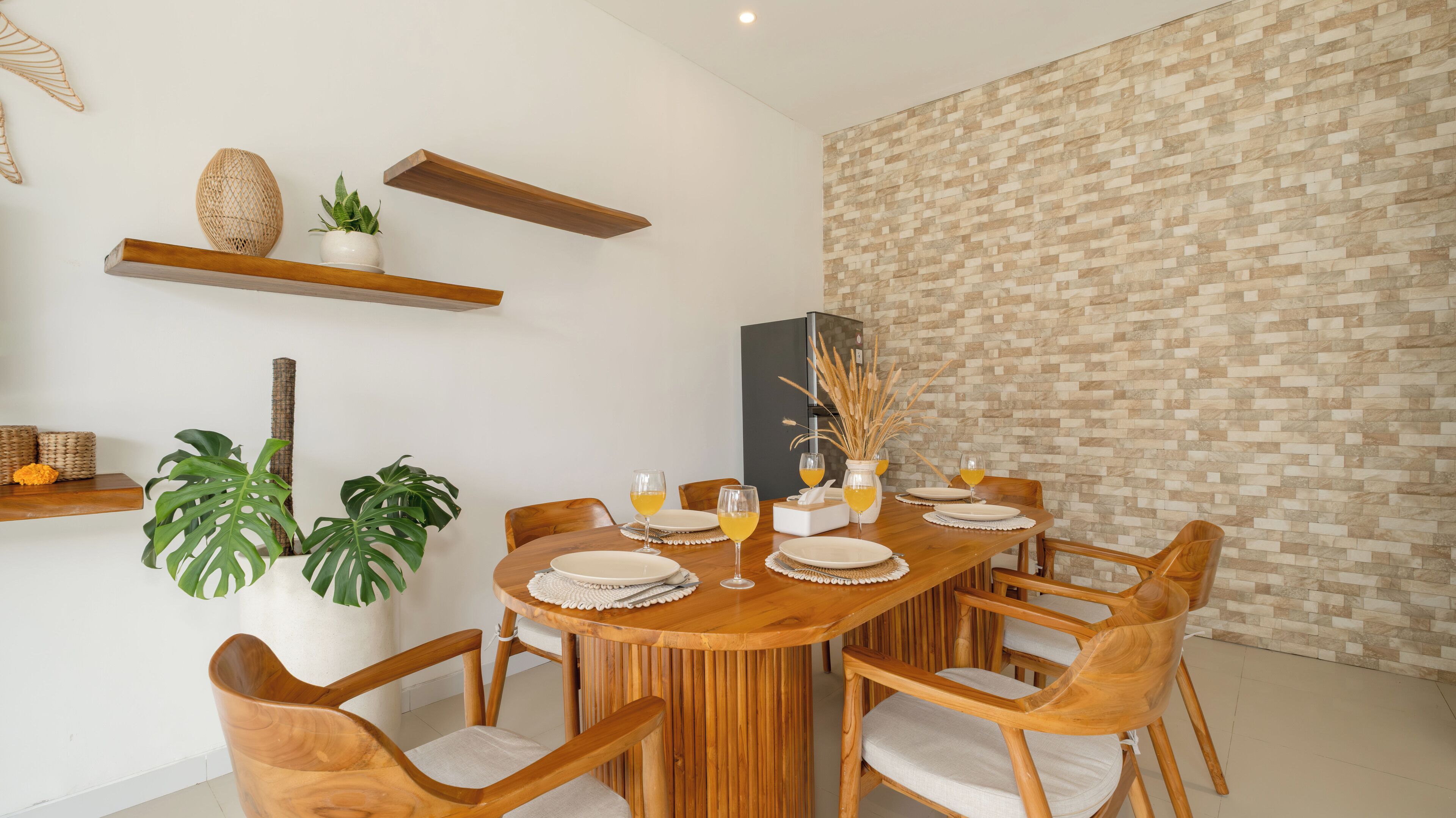 A dining area with a wooden table set for six, surrounded by wooden chairs with cushioned seats. There are two potted plants, one large and one small, and a decorative vase on a shelf above the table.