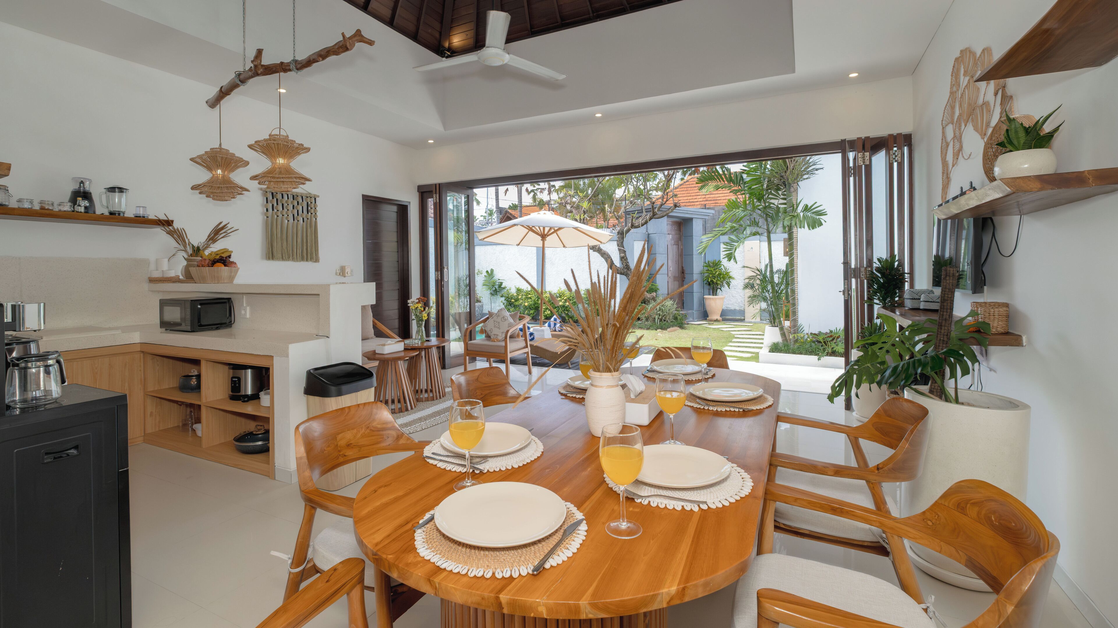 Uma sala de jantar moderna com uma mesa de madeira posta para seis pessoas, cercada por cadeiras de madeira. Há uma área de cozinha à esquerda, com micro-ondas e cafeteira. O quarto possui grandes janelas que oferecem vista para um jardim e um edifício lá fora.