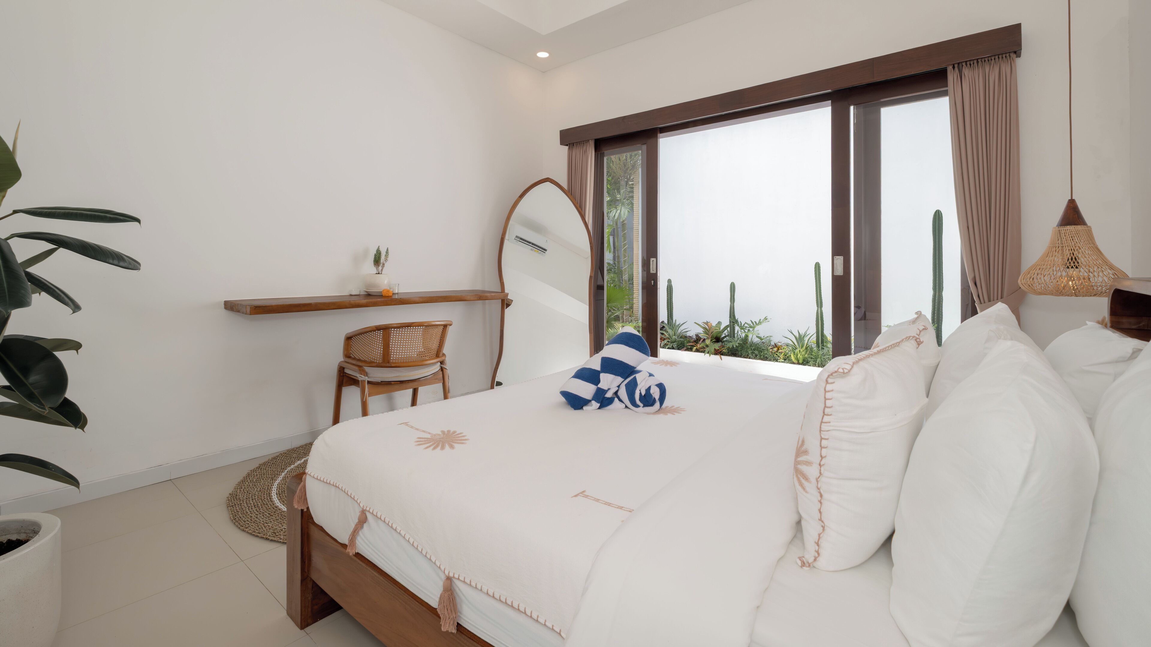 A bedroom with a large bed, a wooden chair, a small table with a plant, and a window with curtains.