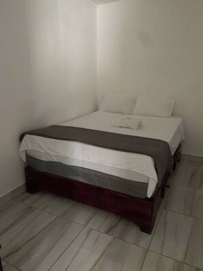 Ein kleines, ordentlich bezogenes Bett in einem Eckzimmer mit weißen Wänden und Fliesenboden.