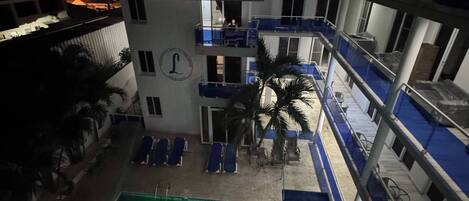 A multi-story building with balconies, a swimming pool with a green logo, and a palm tree.