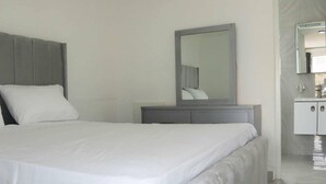 Un dormitorio moderno con una cama grande, un cabecero gris, una mesita de noche con espejo y un baño pequeño con lavamanos y armario.
