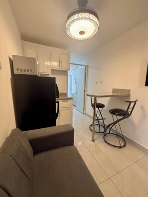 Una cocina compacta con armarios blancos, un refrigerador negro, una mesa de comedor con dos sillas y una lámpara en el techo.