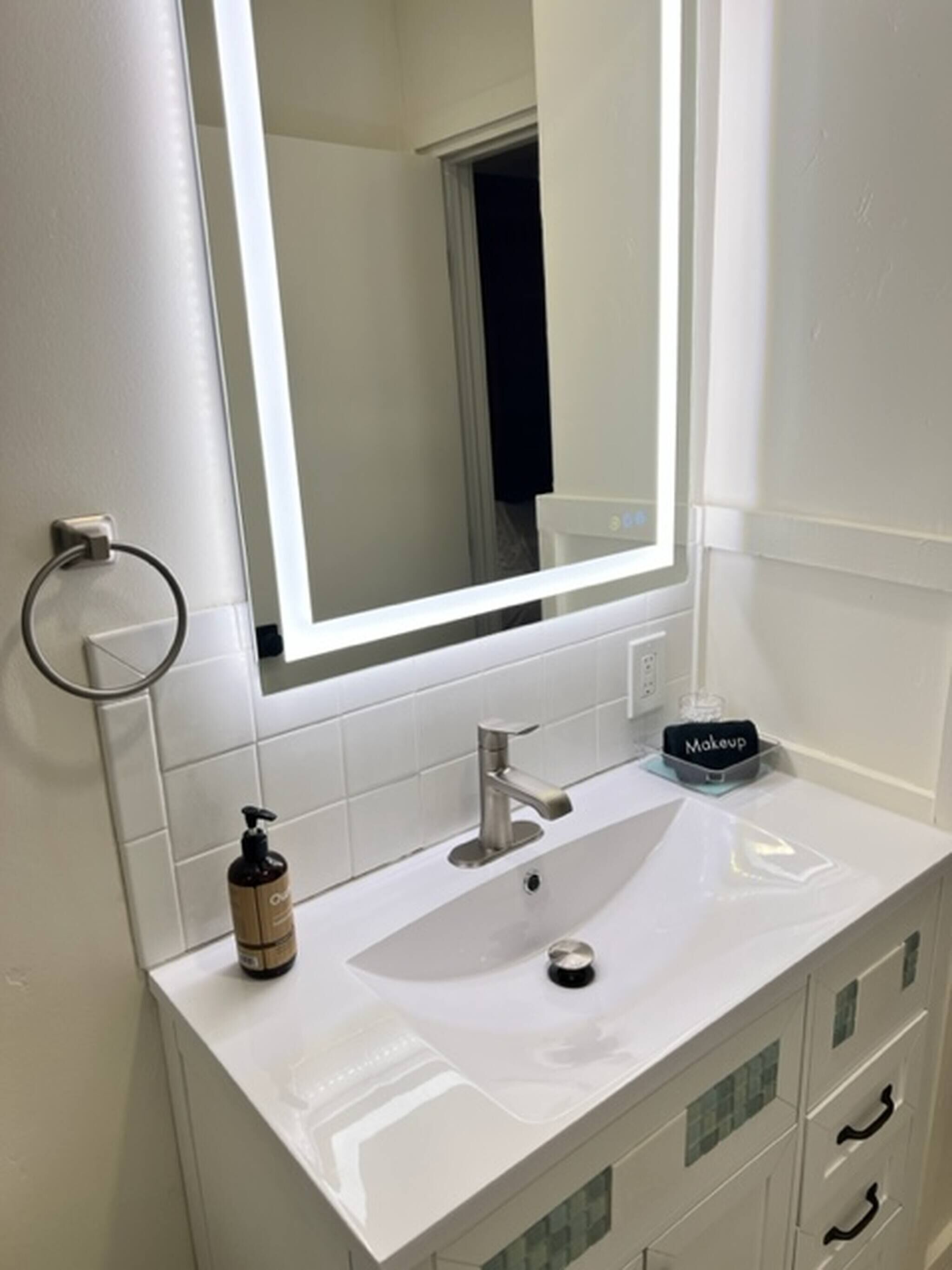 Un baño con un lavamanos blanco, un espejo, un organizador de maquillaje y un dispensador de jabón para manos.