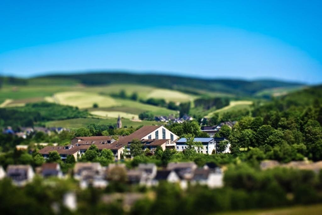 A town with a prominent white building with a steep roof, surrounded by greenery and rolling hills.