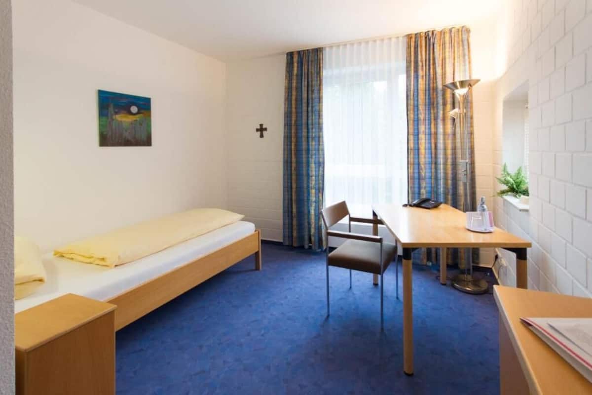 A room with a bed, a desk, a chair, a lamp, and a painting on the wall.