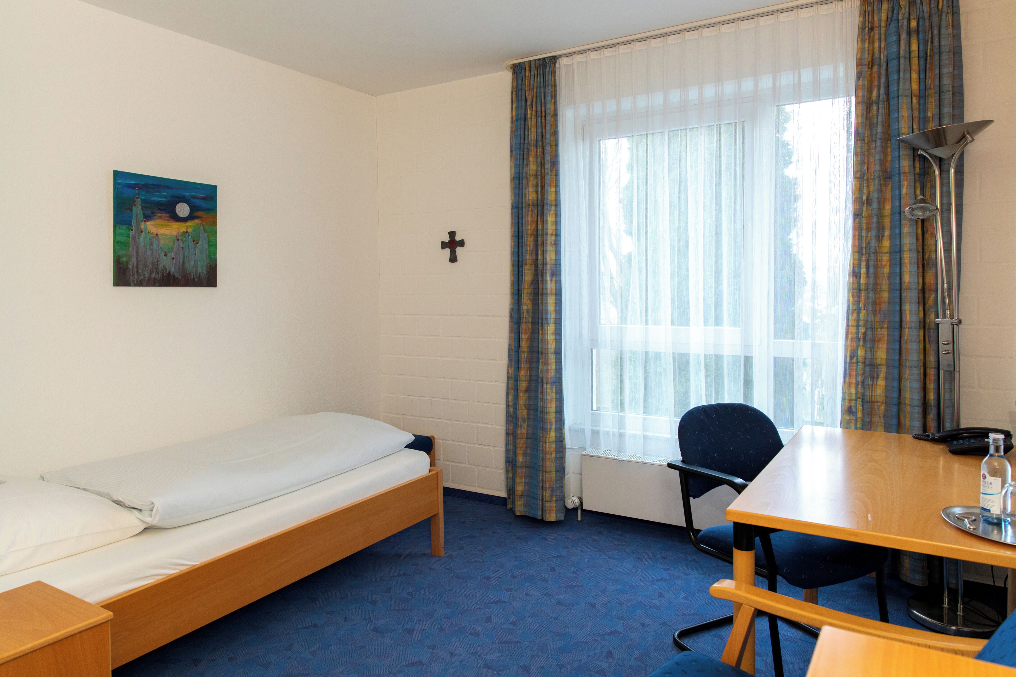 A single bed, a desk with a chair, a painting on the wall, a cross, and a window with curtains.
