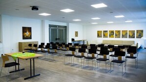 Une salle de conférence avec des rangées de chaises, une table recouverte d’une nappe verte et des œuvres d’art accrochées aux murs.