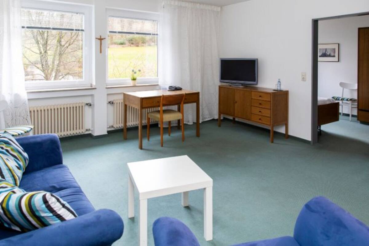 A room with a blue sofa, a white table, a TV on a wooden cabinet, and a window with a view of trees.