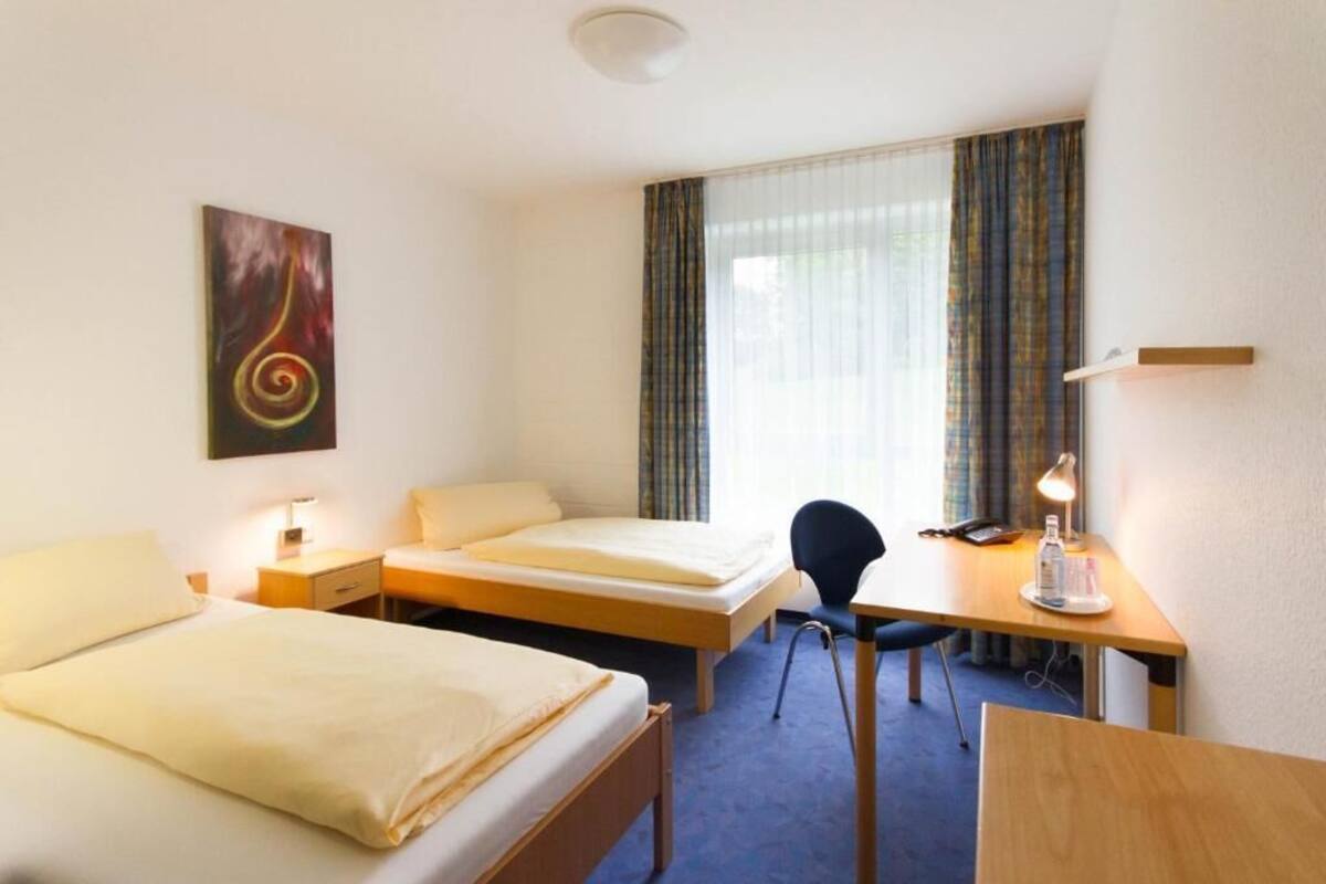 A hotel room with two beds, a desk, a chair, a desk lamp, a telephone, and a painting on the wall.