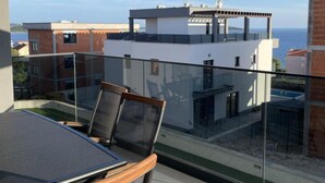 A balcony with a glass railing, a table, and two chairs. In the background, there is a building with a white facade and a view of the sea. - Apartment Rogoznica (Rogoznica)
