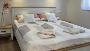 A neatly made bed with a white and gray striped blanket, a folded towel, and several pillows. There is a bedside table with a vase of dried flowers. - Apartment Rogoznica (Rogoznica)